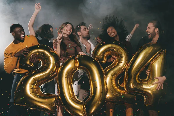 Group of joyful young people celebrating 2024 New Year while dancing and throwing confetti in night club