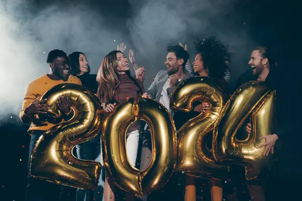 Group of joyful young people holding 2024 gold numbers while celebrating New Year in night club together