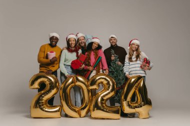Mutlu insanlar 2024 yılını arka planda kutlarken ellerinde Noel süsleri ve hediye kutuları tutuyorlar.