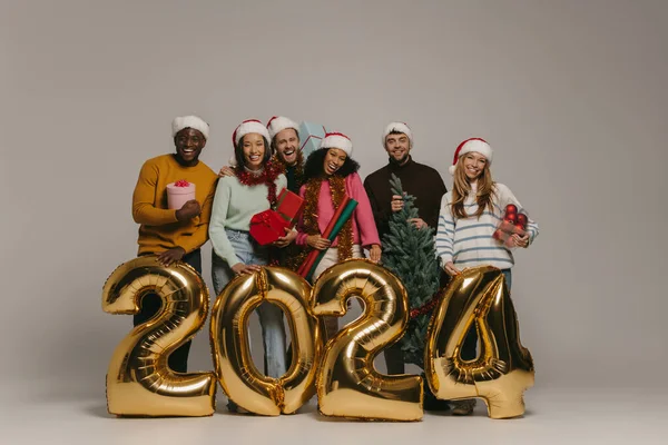 Joyful people holding Christmas ornaments and gift boxes while celebrating 2024 New Year on background