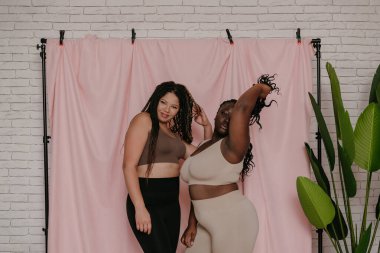 Kumaş arka planda sırıtırken gülümseyen, spor kıyafetli, iri yarı iki Afrikalı kadın.