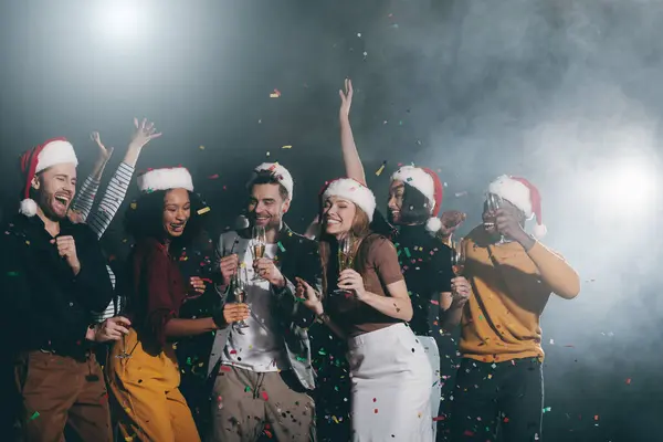 Group of joyful young people celebrating New Year in night club together while confetti flying around