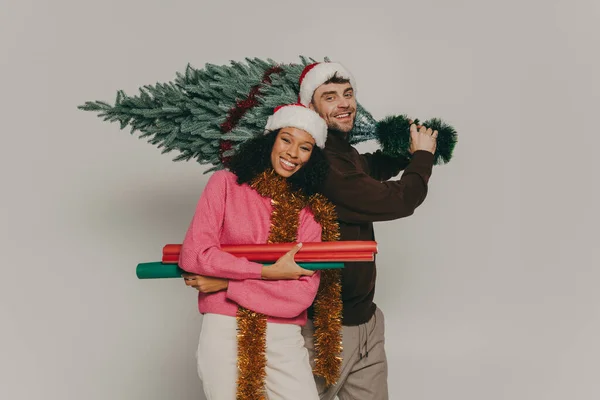 一对戴着圣诞礼帽 头戴圣诞礼帽 头戴圣诞礼帽的年轻夫妇在工作室背景下抱着圣诞树 — 图库照片