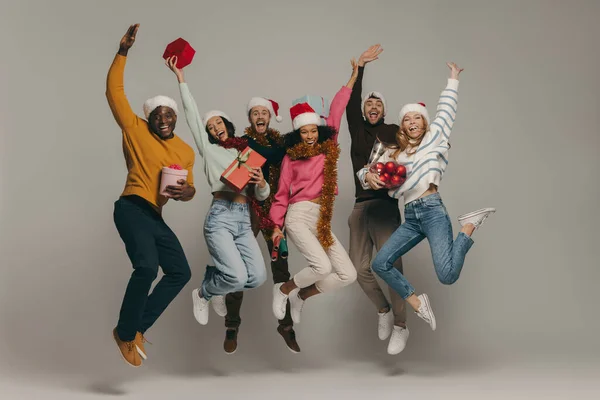 Joyful young people carrying Christmas ornaments and gift boxes while jumping on background together