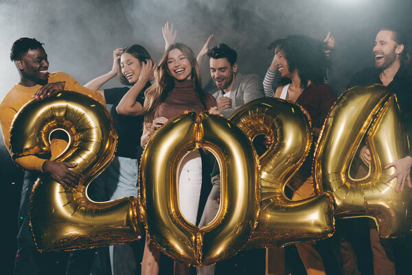 Group of joyful young people holding 2024 shaped gold balloons while celebrating New Year in night club