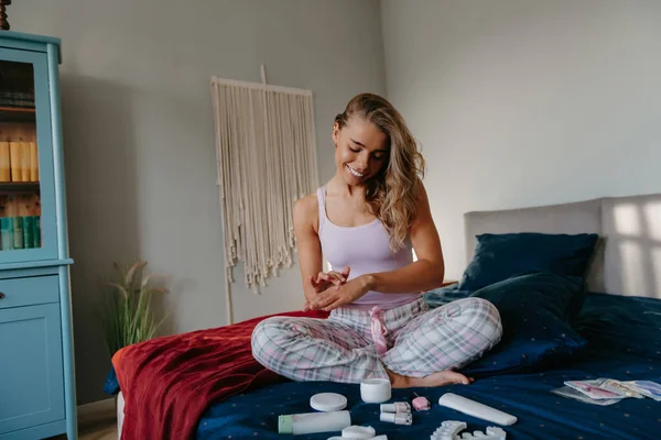 Young woman testing various skin care products while applying hydrating cosmetic cream on hand at home