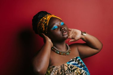 Kendine güveni tam ve güzel makyajlı Afrikalı bir kadın. Kırmızı arka planda dikilirken geleneksel kıyafetler giyiyor.