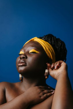 Kendine güvenen, iri yarı, güzel makyajlı, mavi arka planda küpe takan Afrikalı bir kadının portresi.