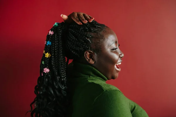 Side view of happy African woman with braided hair and colorful clips standing on red background