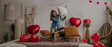 Mutlu genç çift yatakta yastık savaşı yapıyor. Kırmızı kalp şeklindeki balonlarla çevrili.