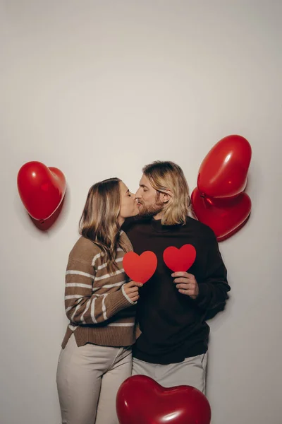 Loving couple holding heart shape Valentines cards and kissing on beige background with balloons flying around