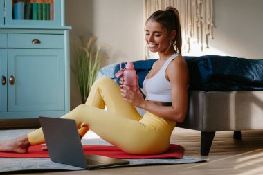 Mutlu sportif kadın su şişesini tutuyor ve evinde egzersiz minderinde dinlenirken dizüstü bilgisayara bakıyor.