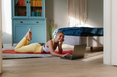 Ev çalışmasından sonra spor paspası üzerinde dinlenirken dizüstü bilgisayar kullanan mutlu bir kadın.