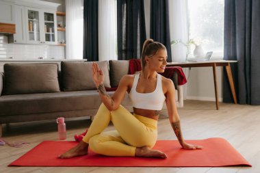 Spor kıyafetleriyle gülümseyen genç bir kadın evde egzersiz minderinde yoga yapıyor.