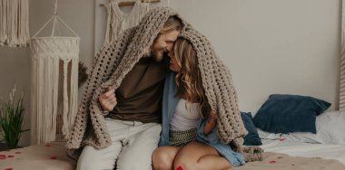 Sıcak bir battaniyeyle örtünen ve yatakta birlikte otururken gülümseyen genç ve mutlu bir çift.