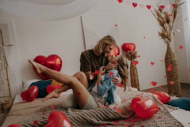 Romantik çift yatakta oturup öpüşürken kırmızı kalp şeklinde konfeti uçuşuyor.