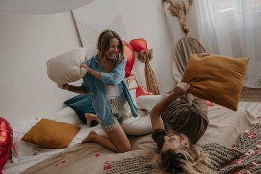 Mutlu genç çift yatakta yastık savaşı yapıyor. Kırmızı kalp şeklinde balonlarla çevrili.