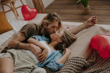 Kırmızı kalp şeklindeki balonlarla çevrili yatakta uzanmış öpüşen romantik çift.