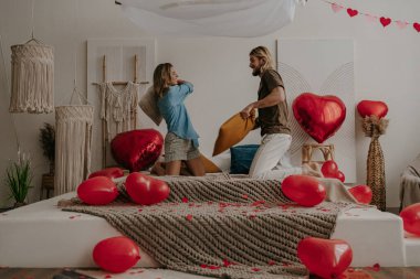 Mutlu genç çift yatakta yastık savaşı yapıyor. Kırmızı kalp şeklindeki balonlarla çevrili.