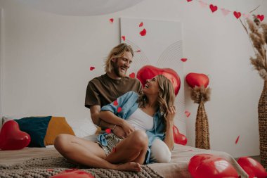 Sevgililer Günü 'nü evde kutlarken yatakta oturup kalp şeklinde konfeti atan neşeli bir çift.