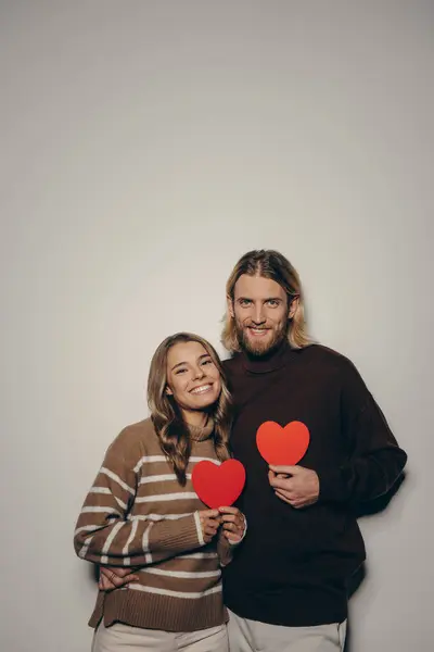 Smiling loving couple holding heart shape Valentines cards while standing on beige background together