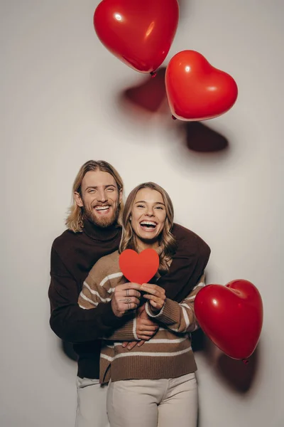 Joyful loving couple holding heart shape Valentine cards on beige background with balloons flying around