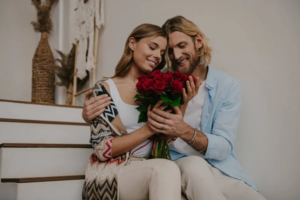 Romantic young man giving a flower bouquet his happy girlfriend while sitting on stairs at home together