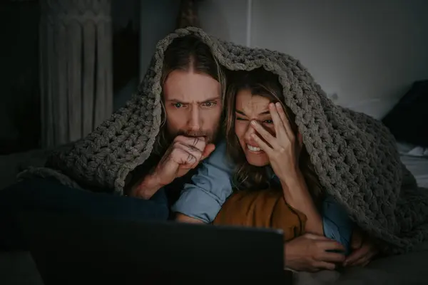 Scared young couple covering with warm blanket and looking at laptop while lying in bed at home together