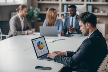 Güvenilir iş adamı, meslektaşları arka planda birlikte çalışırken dizüstü bilgisayardaki grafiği analiz ediyor.