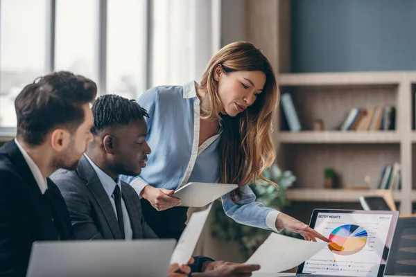 Üç kendine güvenen iş adamı pazarlama verilerini analiz ediyor ve ofiste birlikte çalışırken dizüstü bilgisayar kullanıyor.