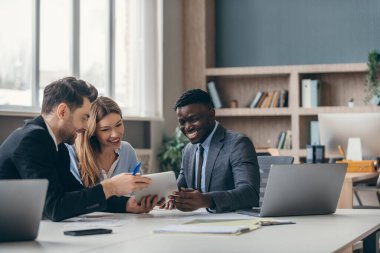 Resmi giyimde üç kendine güvenen insan ofiste iş tartışırken teknolojilerini kullanıyorlar.