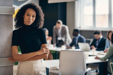 İş arkadaşları arka plan üzerinde çalışırken, kendine güvenen Afrikalı iş kadını kollarını kavuşturuyor.