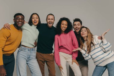 Mutlu genç arkadaşlar bej arka planda birlikte dururken kucaklaşıp gülümsüyorlar.