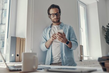 Ofisteki iş yerinin yanında dururken akıllı telefon kullanan kendine güvenen genç adam.