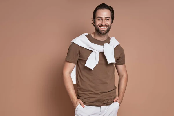 Fashionable young man holding hands in pockets and smiling against brown background