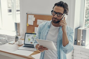 Ofiste dikilirken telefonla konuşan ve elinde dijital tablet tutan kendine güvenen bir adam.