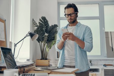 Ofisteki iş yerinin yanında duran kendine güvenen genç adam akıllı bir telefon tutuyor.