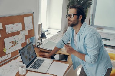 Ofisteki iş yerinin yanında duran yakışıklı genç adam düşünceli görünüyor.