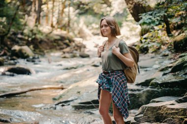 Sırt çantalı, kendinden emin genç bir gezgin dağ nehri boyunca taşların yanında yürüyor.