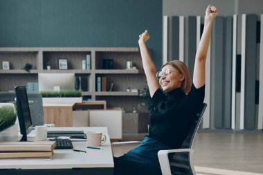 Ofis masasında otururken kollarını açık tutan mutlu genç iş kadını.