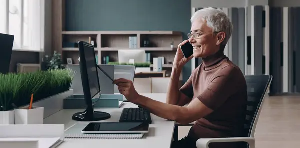 Empresária Sênior Feliz Falando Telefone Apontando Monitor Computador Enquanto Trabalhava Fotografias De Stock Royalty-Free