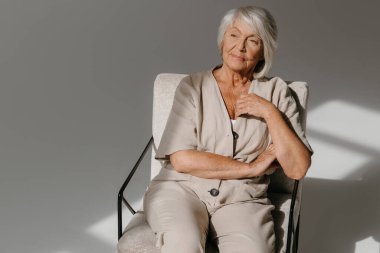 Rahat, gri saçlı, yaşlı bir kadının rahat koltuğunda oturması ve sakin görünmesi.