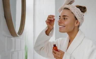 Bornozlu genç bir kadın banyoda dikilirken yüzüne kozmetik serumu sürüyor.