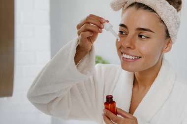 Bornozlu genç bir kadın banyoda dikilirken yüzüne kozmetik serumu sürüyor.