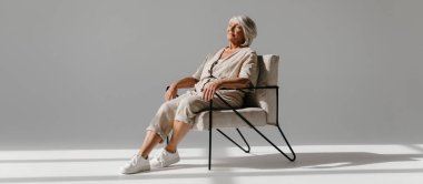 Rahat gri saçlı yaşlı kadın rahat koltukta oturuyor ve arka planda sakin görünüyor.