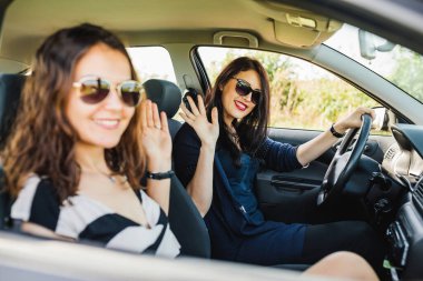 Profil pozu. İki güzel kadın arabada otururken kameraya el kaldırıyor.