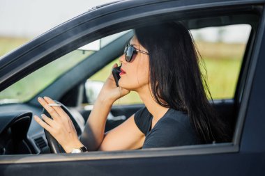 Araba kullanırken sigara içen ve telefonla konuşan genç bir kadının profili.