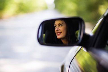 Front view from the car mirror with woman driver smiling reflected on it .