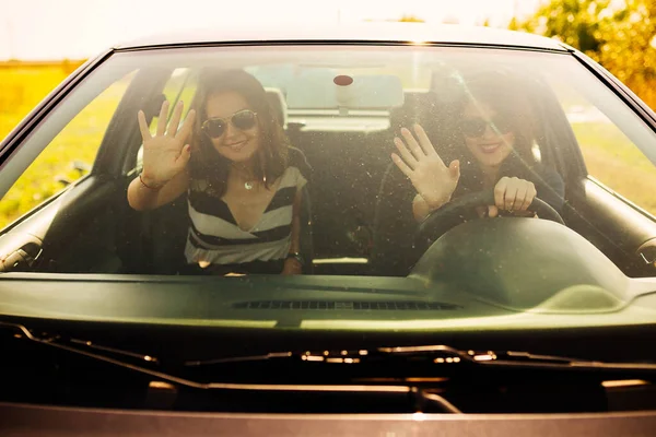 Youth lifestyle - two smiling woman friends rising a hand to the camera and preparing to drive.