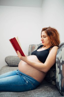 Güzel hamile kadın gündelik ve rahat kıyafetler giyiyor ve kanepede kitap okuyor..
